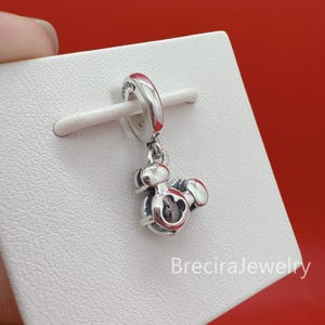 May include: A silver-toned metal charm featuring a stylized Mickey Mouse head silhouette with a central clear stone. The charm has a circular bail engraved with the word Disney and is mounted on a white display card with the text BreciraJewelry.