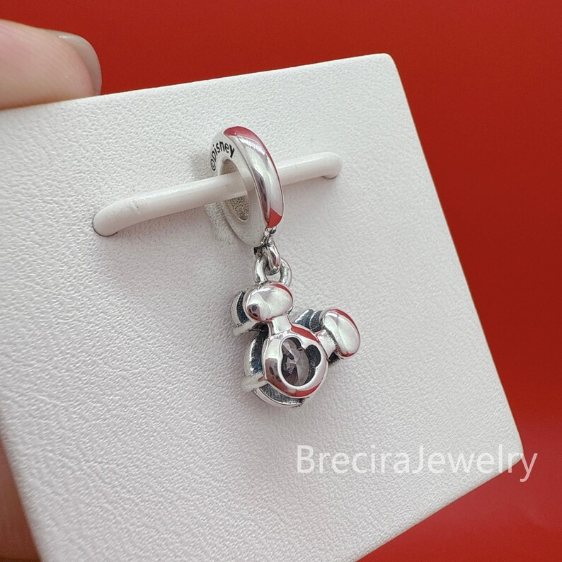 May include: A silver-toned metal charm featuring a stylized Mickey Mouse head silhouette with a central clear stone. The charm has a circular bail engraved with the word Disney and is mounted on a white display card with the text BreciraJewelry.