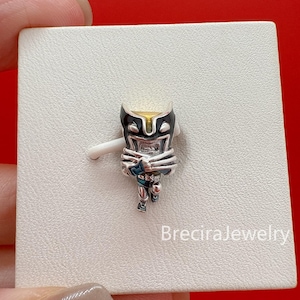 May include: A silver-toned metal charm featuring a stylized character with a yellow forehead, black mask details, and crossed arms. The charm has a polished finish and is attached to a white display card with the text BreciraJewelry printed in white.