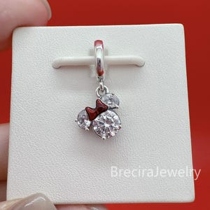 May include: A silver-toned charm pendant featuring a mouse-shaped silhouette adorned with three clear, round-cut stones and a small, glossy red bow. The charm is attached to a silver-toned bail and is displayed on a white textured card.