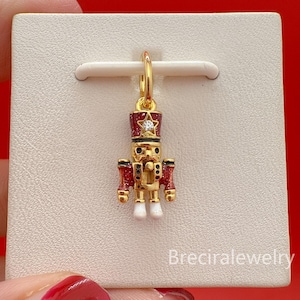 May include: A gold-coloured charm in the shape of a nutcracker soldier. The charm features a red and black hat with a star, red arms, and white boots. It hangs from a gold hoop. The text "Breciralejewelry" is visible.
