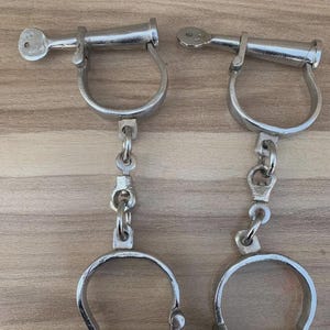 Puede incluir: Dos esposas de metal plateado con llaves y cadenas de conexión. Las esposas están hechas de metal grueso y redondeado y están diseñadas para asegurar las muñecas. Las llaves se insertan en el mecanismo de bloqueo. Las esposas se muestran sobre una superficie de madera marrón claro.