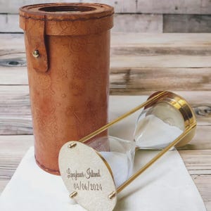 Puede incluir: Un reloj de arena con marco de latón y arena blanca, junto a un estuche de cuero marrón. El reloj de arena tiene una base dorada con el texto "Langkawi Island 04/06/2024". El estuche es cilíndrico y decorado con símbolos náuticos.