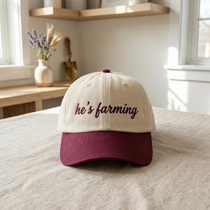 May include: Cream-colored baseball cap with a maroon brim and top button. The phrase "he's farming" is embroidered in maroon script across the front. The hat sits on a neutral-toned surface.