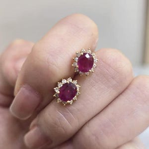 Könnte beinhalten: Ein Paar goldene Ohrstecker, jeweils mit einem großen ovalen Rubin, der von einem Kranz aus kleinen, funkelnden Diamanten umgeben ist. Die Ohrringe sind in einer Goldfassung gehalten und erzeugen einen klassischen und eleganten Look.