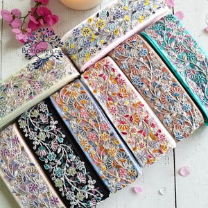 May include: A collection of floral embroidered ribbons in various colors, including white, pink, black, and blue. The ribbons are adorned with delicate floral patterns and are arranged on a white wooden surface. A lit candle and pink flowers are in the background.