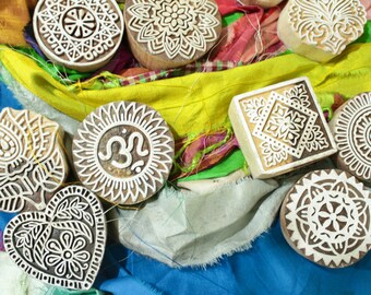 May include: A collection of wooden printing blocks with intricate designs, including a lotus flower, an Om symbol, a heart, and various geometric patterns. The blocks are arranged on a colorful fabric background.