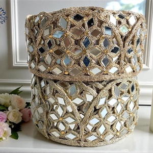 Puede incluir: Dos recipientes cilíndricos dorados con un diseño de mosaico de espejo. Los recipientes tienen un borde tejido y con cuentas. Las superficies espejadas reflejan la luz. Una vela y flores están en el fondo. Decoración del hogar.