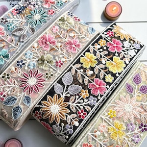 Puede incluir: Cuatro rollos de cinta decorativa de tela con intrincados bordados florales sobre malla transparente. Cada rollo muestra una paleta de colores única, incluyendo bases blancas, beige, negras y verde claro con patrones de flores multicolores.