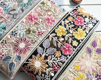 Puede incluir: Cuatro rollos de cinta decorativa de tela con intrincados bordados florales sobre malla transparente. Cada rollo muestra una paleta de colores única, incluyendo bases blancas, beige, negras y verde claro con patrones de flores multicolores.