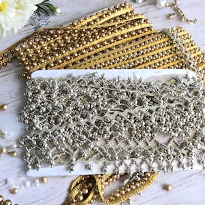 Op de afbeelding: Twee spoelen met parelversiering, een gouden en een zilveren. De gouden versiering heeft ronde parels en een metallic afwerking. De zilveren versiering heeft een ingewikkelder ontwerp met kleinere parels en een meer delicate uitstraling.