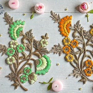 Peut inclure: Deux appliques florales brodées, une verte et une orange, avec des accents dorés et argentés. Les appliques sont ornées de paillettes et sont entourées de petites bougies roses et de roses.