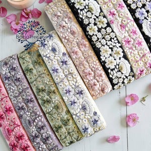 May include: A collection of embroidered floral trim ribbons in various colors, including pink, purple, green, and black. Each ribbon features detailed floral patterns with white and silver accents. The ribbons are arranged on a white wooden surface with scattered pink flower petals.