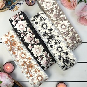 May include: Three rolls of decorative fabric trim featuring intricate floral embroidery. One roll has a white base with copper-toned flowers, one has a black base with pink and white flowers, and one has a white base with black and white flowers.