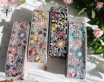 May include: Four different trims of embroidered floral lace ribbon. The trims are in shades of pink, green, black, and blue. The trims are all on a white background.