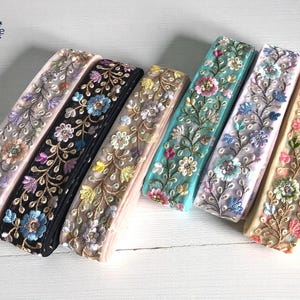 Könnte beinhalten: Eine Sammlung von bestickten Blumenbändern in verschiedenen Farben. Die Bänder weisen detaillierte Blumenmuster auf, darunter Schwarz, Türkis, Rosa und Creme. Die Bänder sind nebeneinander angeordnet und zeigen die verschiedenen Designs.