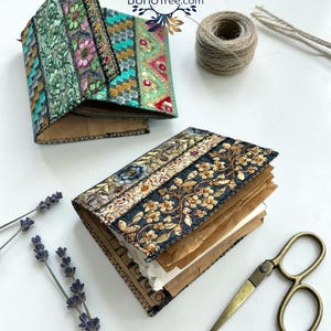K&ouml;nnte beinhalten: Zwei kleine Notizb&uuml;cher mit Stoffeinb&auml;nden, die mit aufwendigen Blumenstickereien in Gr&uuml;n-, Gold- und Schwarzt&ouml;nen verziert sind. Die Seiten bestehen aus braunem Papier. Ein Notizbuch ist ge&ouml;ffnet, das andere geschlossen. Das Logo BohoTree.com ist sichtbar.