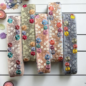 May include: Five rolls of decorative fabric trim featuring intricate floral embroidery on a sheer mesh base. Each roll displays a unique color palette including beige, sage green, peach, white, and grey, adorned with multi-colored thread flowers.