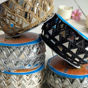 May include: Decorative trim rolls with mirrored triangle accents in gold, silver, and black. The trim is displayed on spools with blue and orange labels. The trim is suitable for various craft and design projects.
