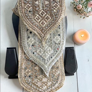 May include: An ornate, beige and silver scarf with intricate embroidery and mirrored accents. The scarf is displayed on a black mannequin head, with a lit candle and flowers in the background. The scarf has a detailed, geometric pattern.
