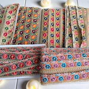 May include: Various rolls of decorative floral trim in multiple colors. The trim features embroidered flowers in red, pink, blue, and orange, with gold and beige accents. The trim is likely used for embellishing clothing or crafts.