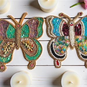 May include: Two decorative butterfly ornaments. One is green and purple, the other is blue and rainbow-colored. Both are embellished with sequins and beads, with gold-colored edging. The butterflies are on a white wooden surface, with candles and flowers.