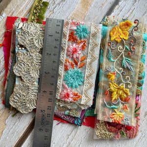 May include: Assortment of colorful embroidered fabric trims with floral and geometric patterns. The trims are layered on top of each other and include a ruler for scale.