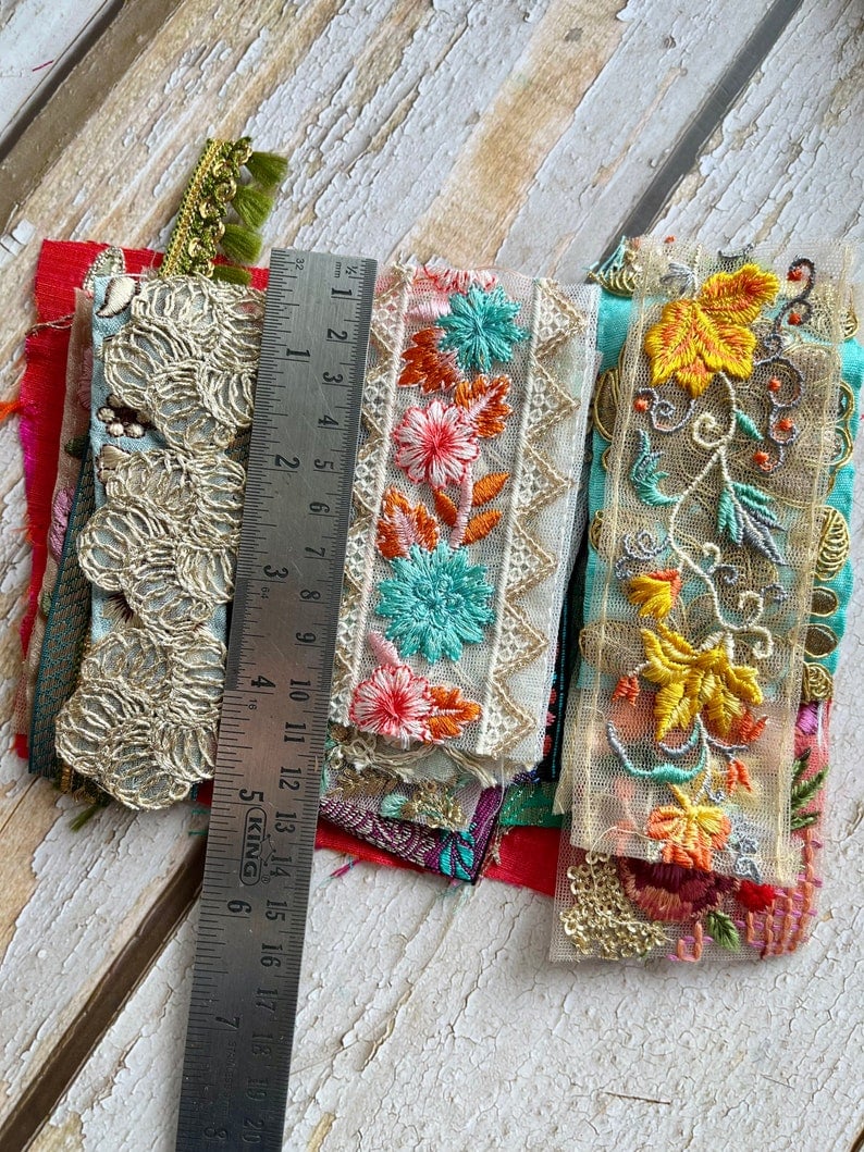May include: Assortment of colorful embroidered fabric trims with floral and geometric patterns. The trims are layered on top of each other and include a ruler for scale.