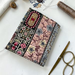 K&ouml;nnte beinhalten: Ein rechteckiger Notizbuchumschlag aus Stoff mit Patchwork-Design und floraler Stickerei in Schwarz, Rosa und Grau. Der Umschlag zeigt komplexe Stickmuster, darunter ein schwarzes Mittelteil mit bunten Blumen und einen gesteppten rosa Bereich. Der Text lautet Design 3.