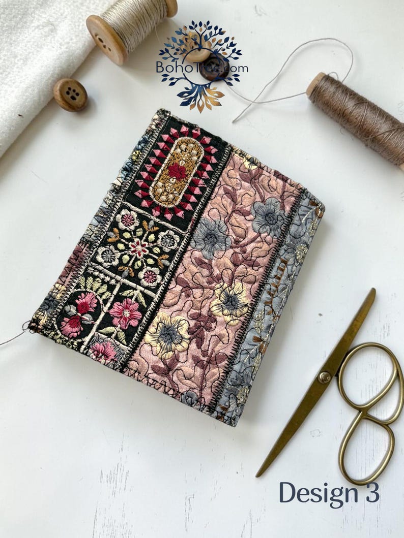 K&ouml;nnte beinhalten: Ein rechteckiger Notizbuchumschlag aus Stoff mit Patchwork-Design und floraler Stickerei in Schwarz, Rosa und Grau. Der Umschlag zeigt komplexe Stickmuster, darunter ein schwarzes Mittelteil mit bunten Blumen und einen gesteppten rosa Bereich. Der Text lautet Design 3.