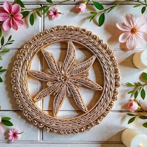 May include: A gold-colored, woven decorative piece with a starburst design in the center. The piece is surrounded by a beaded border. Pink flowers and green foliage are arranged around the piece, along with two white candles.