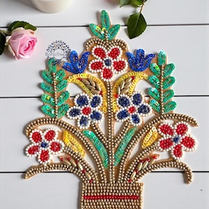 May include: A beaded floral applique in a vase shape. The design features red, blue, and white flowers, green sequined leaves, and gold beads. A red stripe accents the base. The applique is on a white wooden surface.