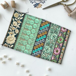 K&ouml;nnte beinhalten: Ein rechteckiger Stoffaufn&auml;her mit vertikalen Bahnen aus aufwendigen Stickereien in Dunkelgr&uuml;n, Mint, Gold und Pink, verziert mit Pailletten und Blumenmotiven. Der Aufn&auml;her ist zusammen mit Holzspulen, einer Schere und Perlen abgebildet.