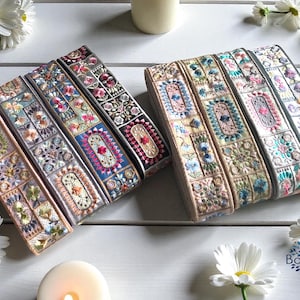 May include: Embroidered ribbon rolls in various colors, featuring floral and geometric patterns. The ribbons are arranged on a white surface with daisies and candles, showcasing a bohemian aesthetic.