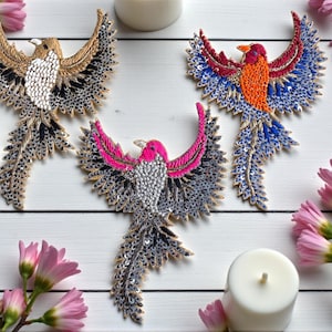 Puede incluir: Tres parches decorativos de pájaros con las alas extendidas, adornados con lentejuelas y abalorios. Los pájaros presentan colores dorados, marrones, rosas, rojos, naranjas y azules. Los parches están dispuestos sobre una superficie de madera blanca con velas y flores rosas.