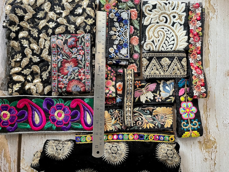 May include: Assortment of colorful embroidered trim with floral and paisley patterns. The trim is on a black background and includes gold and silver accents. A ruler is included for scale.
