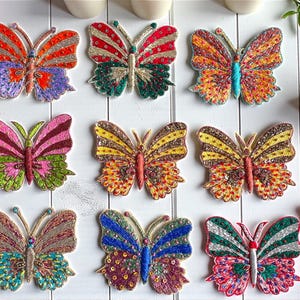 May include: Nine embroidered butterfly patches in various colors, including red, orange, yellow, pink, and blue. Each butterfly features intricate beadwork and detailed wing patterns. The patches are arranged on a white wooden surface.