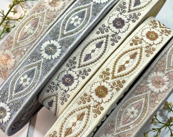 May include: Close-up of several decorative ribbons with floral and geometric embroidered designs. The ribbons feature various colors, including cream, gray, and pink, with gold and silver accents. The ribbons are arranged diagonally, showcasing their intricate details.