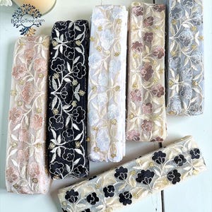 May include: Assortment of embroidered lace ribbons in various colors, including blush, black, white, and gray. Each ribbon features floral patterns with gold accents. The ribbons are arranged on a light-colored surface, showcasing their intricate designs.