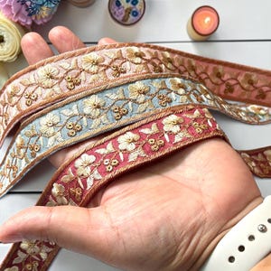 May include: Three strips of decorative fabric trim featuring intricate gold floral embroidery. The ribbons are colored in soft pink, light blue, and deep red, each showcasing a repeating pattern of flowers and leaves with metallic gold thread accents.