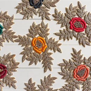May include: Close-up of several embroidered floral appliques. Each applique features a central flower in various colours, including red, orange, and green, surrounded by gold-coloured leaf-shaped embellishments. The appliques are arranged on a white surface.