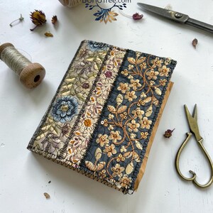 K&ouml;nnte beinhalten: Ein rechteckiges, stoffbezogenes Notizbuch mit einem Patchwork-Design aus drei vertikalen Bahnen. Die Bahnen zeigen aufwendige Blumenstickereien in Gold-, Blau- und Graut&ouml;nen auf strukturiertem Stoff. Der Text Design 3 ist sichtbar.