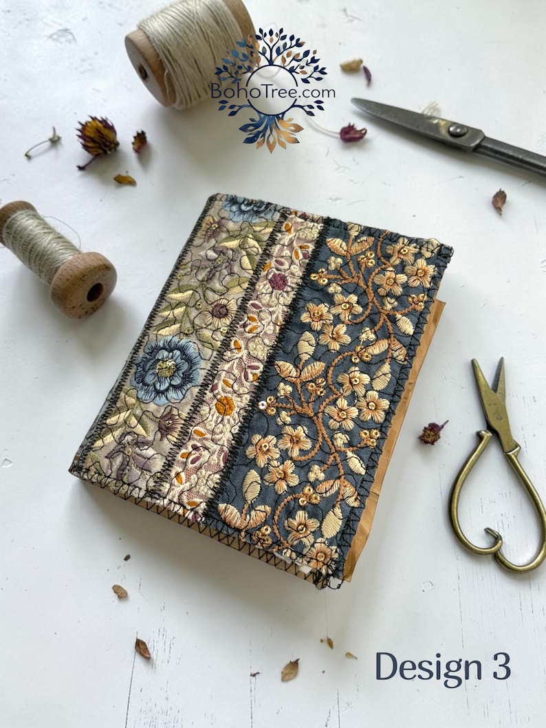 K&ouml;nnte beinhalten: Ein rechteckiges, stoffbezogenes Notizbuch mit einem Patchwork-Design aus drei vertikalen Bahnen. Die Bahnen zeigen aufwendige Blumenstickereien in Gold-, Blau- und Graut&ouml;nen auf strukturiertem Stoff. Der Text Design 3 ist sichtbar.