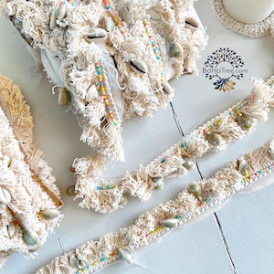 May include: Cream-colored fringe trim with seashells, colorful beads, and a BohoTree.com logo. The trim features a textured, woven design with dangling fringe and is adorned with natural seashells and small, colorful beads. The trim is displayed on a white wooden surface.