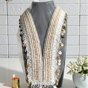Op de afbeelding: Een wit en goud kralen kraagketting met een V-hals ontwerp. De ketting heeft rijen witte kralen en gouden accenten, met gouden muntvormige versieringen die aan de randen hangen. Het woord "Gold" is zichtbaar in de afbeelding.