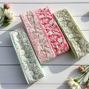 Könnte beinhalten: Vier bestickte Stoffbänder in Pastellfarben: Hellblau, Rosa, Rot und Grün. Jedes Band hat ein weißes Blumenmuster. Die Bänder liegen auf einer weißen Holzoberfläche, mit Blumen und Kerzen.
