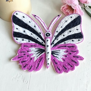 May include: Embroidered butterfly with purple, black, and white wings. The butterfly has a detailed design with striped patterns and dotted accents. The word "Purple" is at the bottom. The butterfly is likely a decorative item or embellishment.