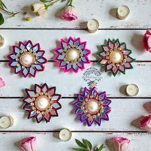 May include: Six decorative, flower-shaped ornaments with mirrored accents and beaded centers. The ornaments are in various colors, including pink, purple, and green, and are arranged on a white wooden surface with candles and flowers.