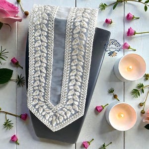 Op de afbeelding: Een wit, ingewikkeld geborduurde kanten kraag met een bladerenpatroon wordt getoond op een donkergrijze vorm. Roze rozen en knoppen, samen met brandende kaarsen, zijn rond de kraag gerangschikt op een wit houten oppervlak.