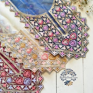 May include: Close-up of intricately embroidered fabric pieces in shades of blue, burgundy, and cream. The designs feature floral patterns with pink, purple, and gold accents. The edges are scalloped, and the fabric appears to be sheer.